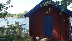 Pieni puinen mökki, jossa on sininen ovi, sijaitsee vesistön rannalla, ja taustalla näkyy laituri.