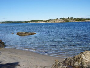 A beach with clear water, a rocky outcrop, and a distant forested hill. - 6 Person Holiday Home in Stromstad (Stromstad)