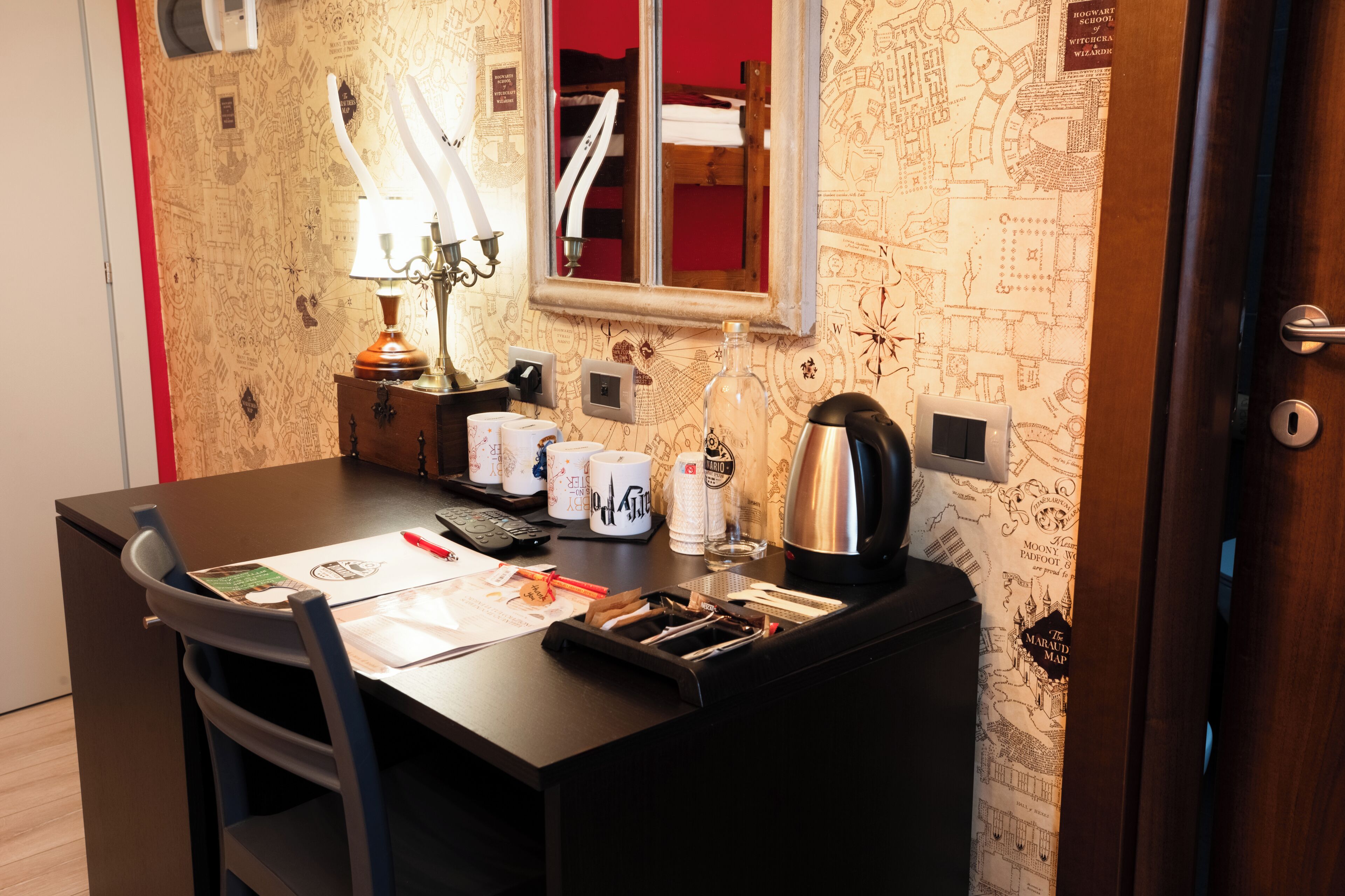 Habitación de hotel con escritorio, lámpara, espejo, papel tapiz con un mapa y una puerta.