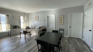 Amplia sala de estar con una gran mesa de comedor, un sofá y una mesa de centro. Hay dos puertas y una ventana con cortinas.