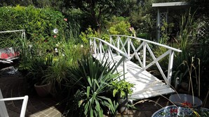 Un pont en bois peint en blanc enjambe un chemin pavé de briques, et est entouré d’une végétation luxuriante et de plantes en pot.