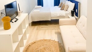 A modern, well-lit room with a bed, a white sofa, a small desk with a TV, and a large woven rug. - Charming studio in center (Cluj-Napoca)