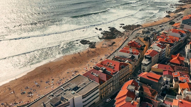 Vista aérea de un pueblo costero con playa, edificios con tejados rojos y una carretera que discurre paralela a la orilla.