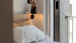 Una cama con sábanas blancas, un cojín color beige y una lámpara de pared con base de madera y pantalla negra.
