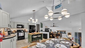 Uma cozinha moderna com armários brancos, uma área de jantar com uma mesa grande posta para seis pessoas e um candelabro com várias luzes esféricas.
