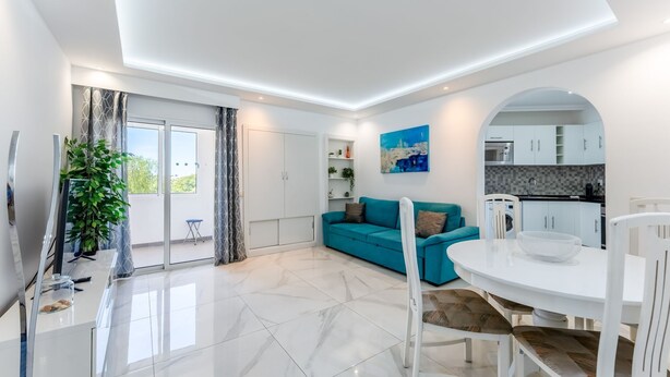 Ein modernes Wohnzimmer mit einem türkisfarbenen Sofa, einem Esstisch mit Stühlen, einer Küchenzeile und einem Balkon mit einem Blumentopf.