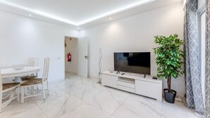 Ein modernes Wohnzimmer mit Flachbildfernseher, Esstisch mit Stühlen, einer Zimmerpflanze und einem Feuerlöscher.