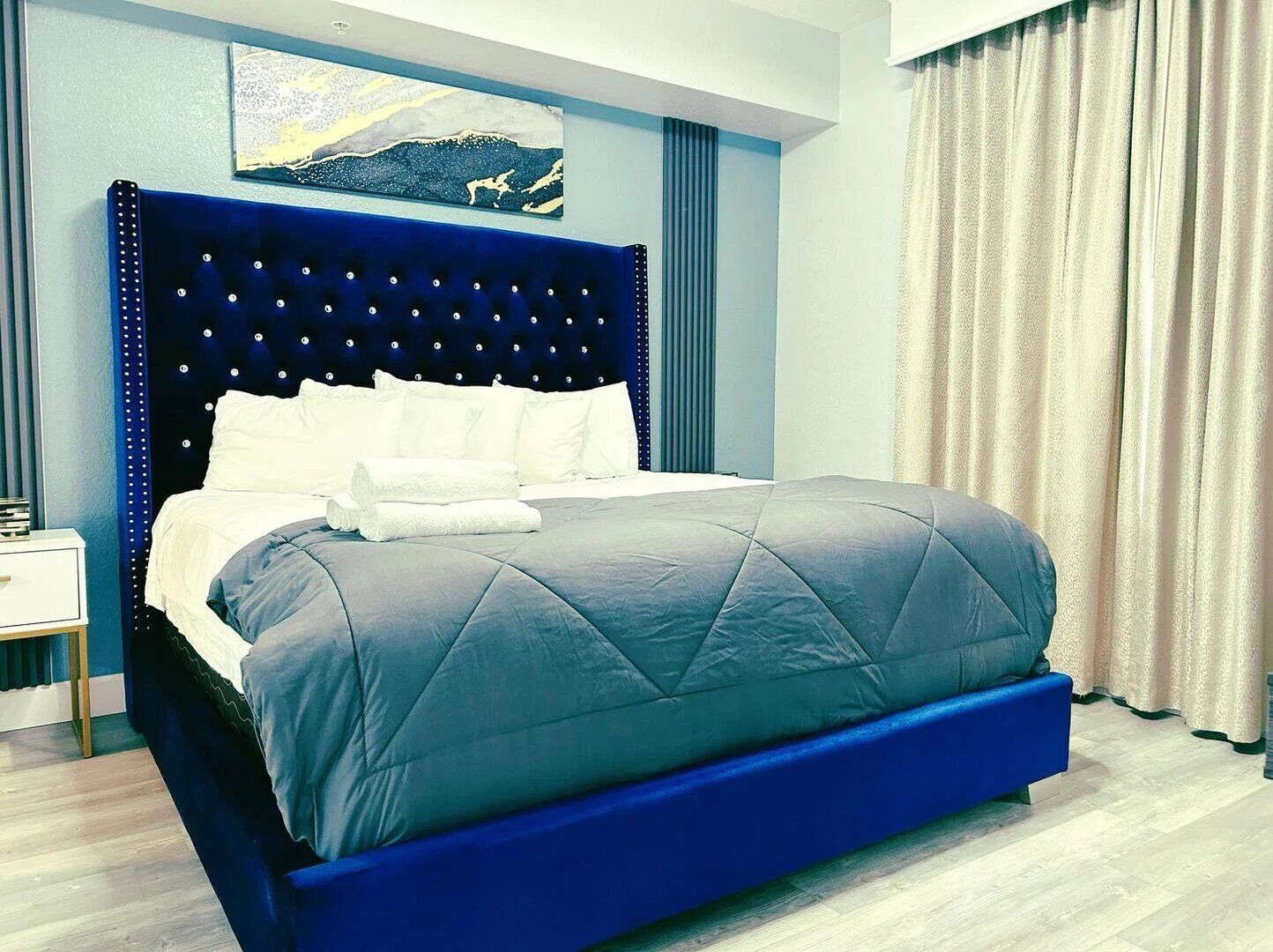 A modern bedroom with a large bed, a blue headboard, and a painting above the bed.