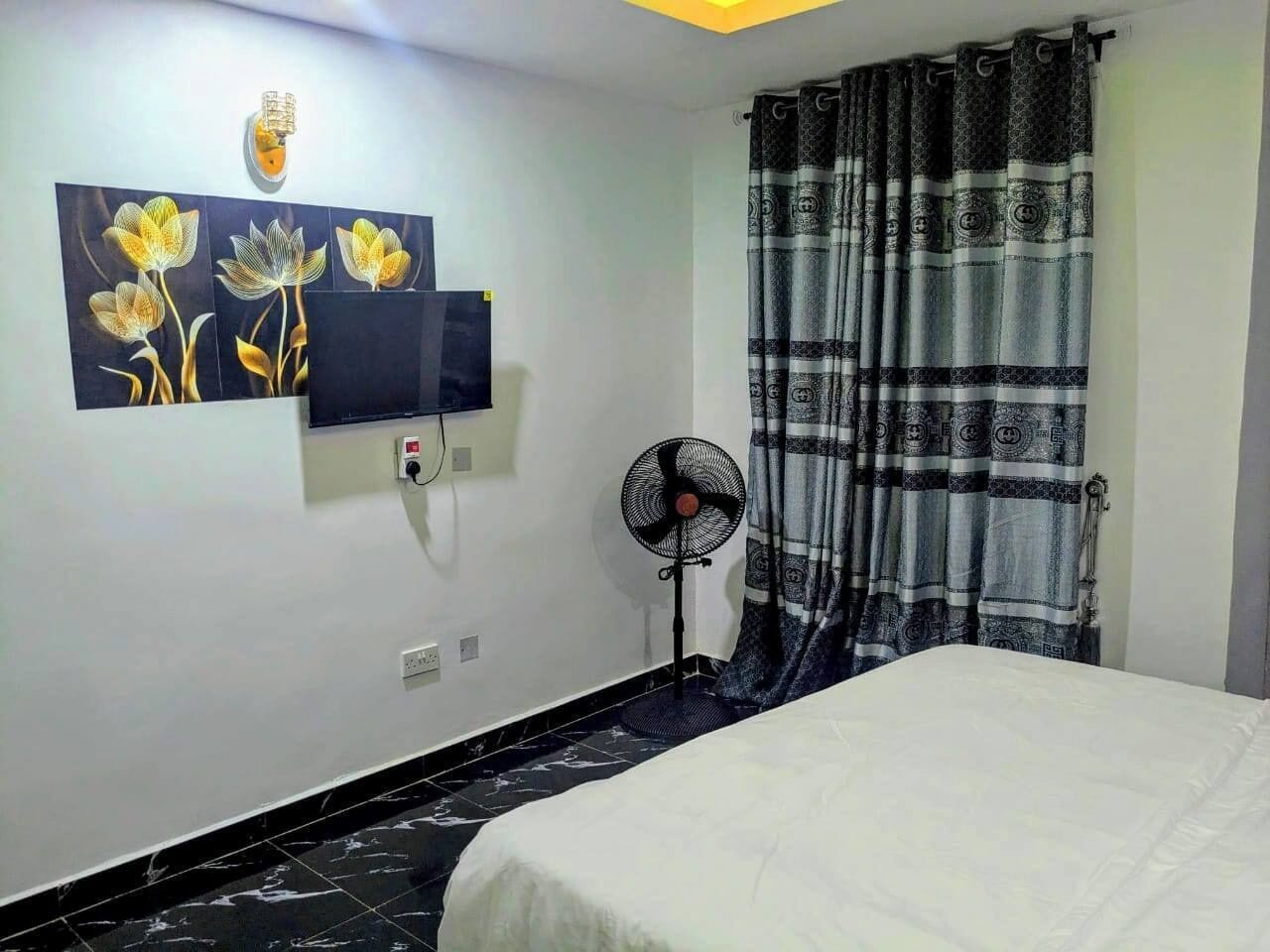 Ein Schlafzimmer mit einem Bett, einem Wand-Fernseher, einem Standventilator und einem gemusterten Vorhang.