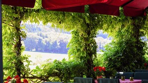 Un área de comedor al aire libre con mesas y sillas, rodeada de abundante vegetación y sombrillas rojas.