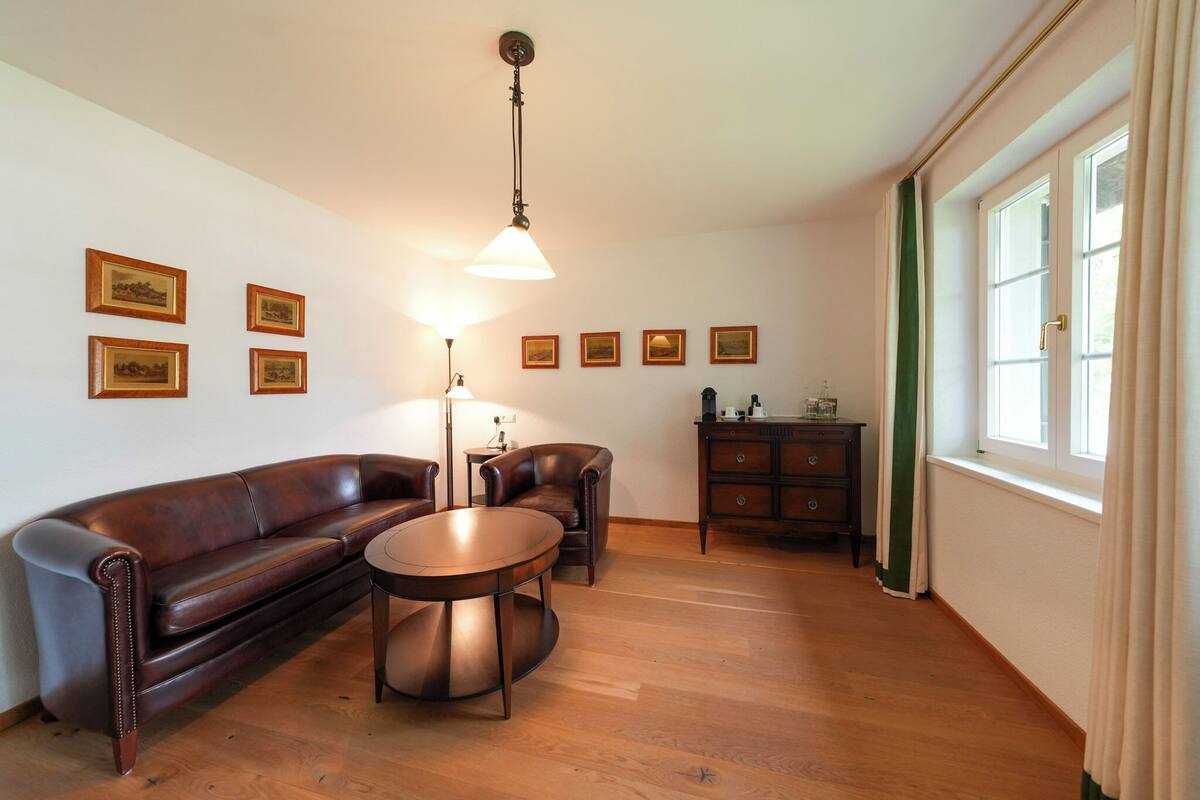 Una sala de estar con un sofá de cuero marrón, una mesa redonda y una araña. Hay cuadros enmarcados en la pared y un ventanal con cortinas.
