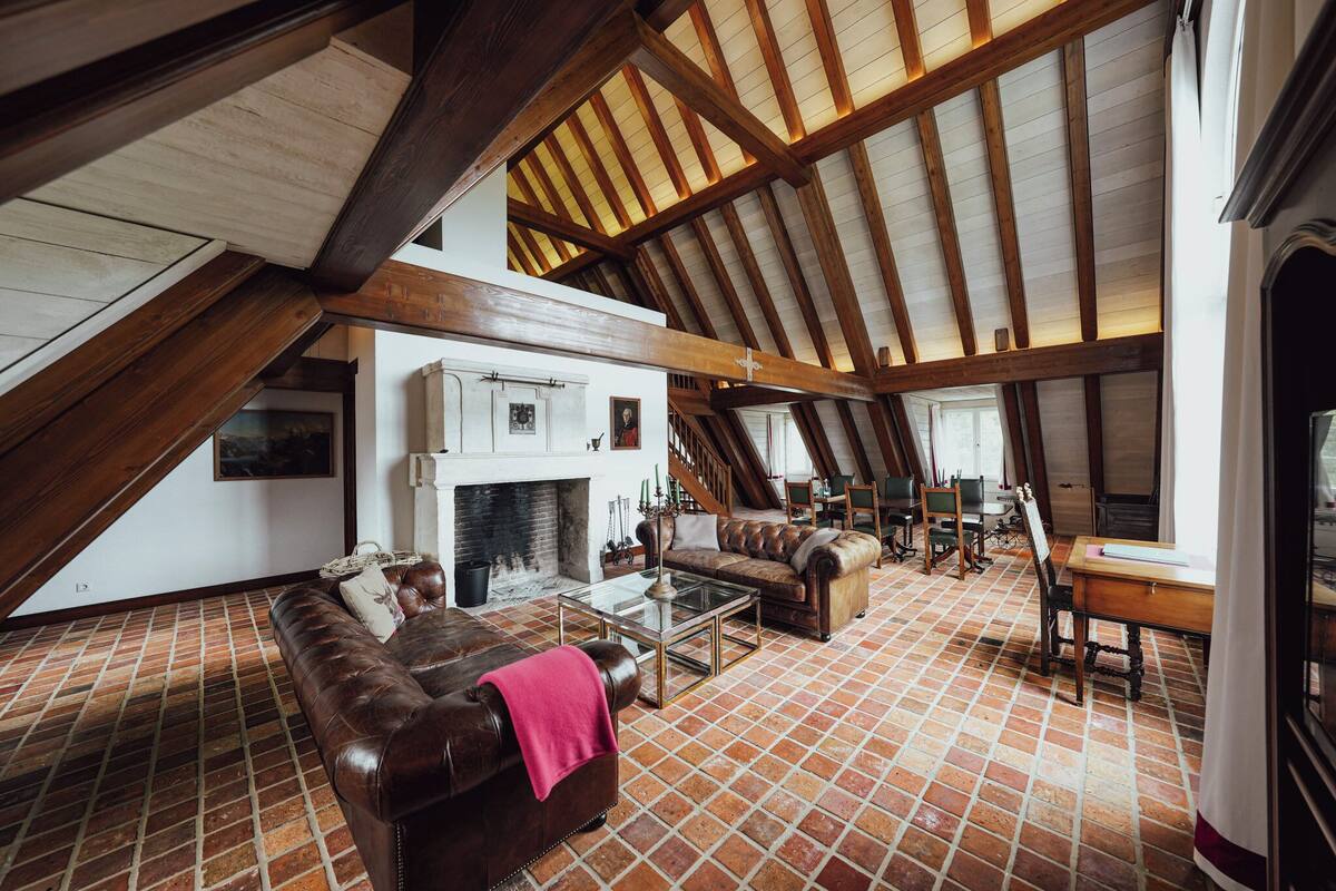 Habitación en ático con vigas de madera a la vista, chimenea, muebles de cuero y suelo de baldosas.