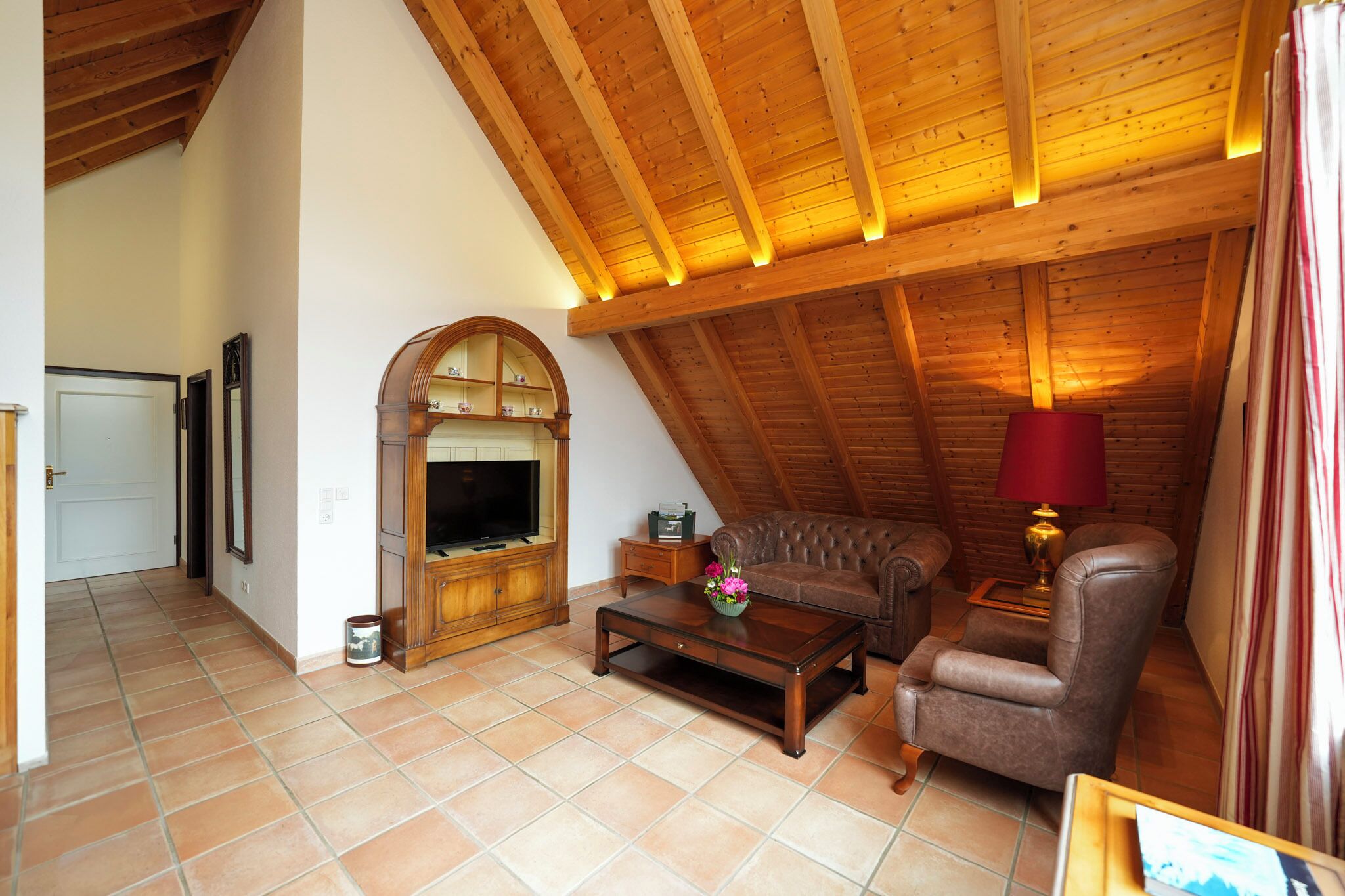 Una sala de estar estilo loft acogedora, con techo de madera, chimenea, televisor, sillón de cuero y una lámpara roja.