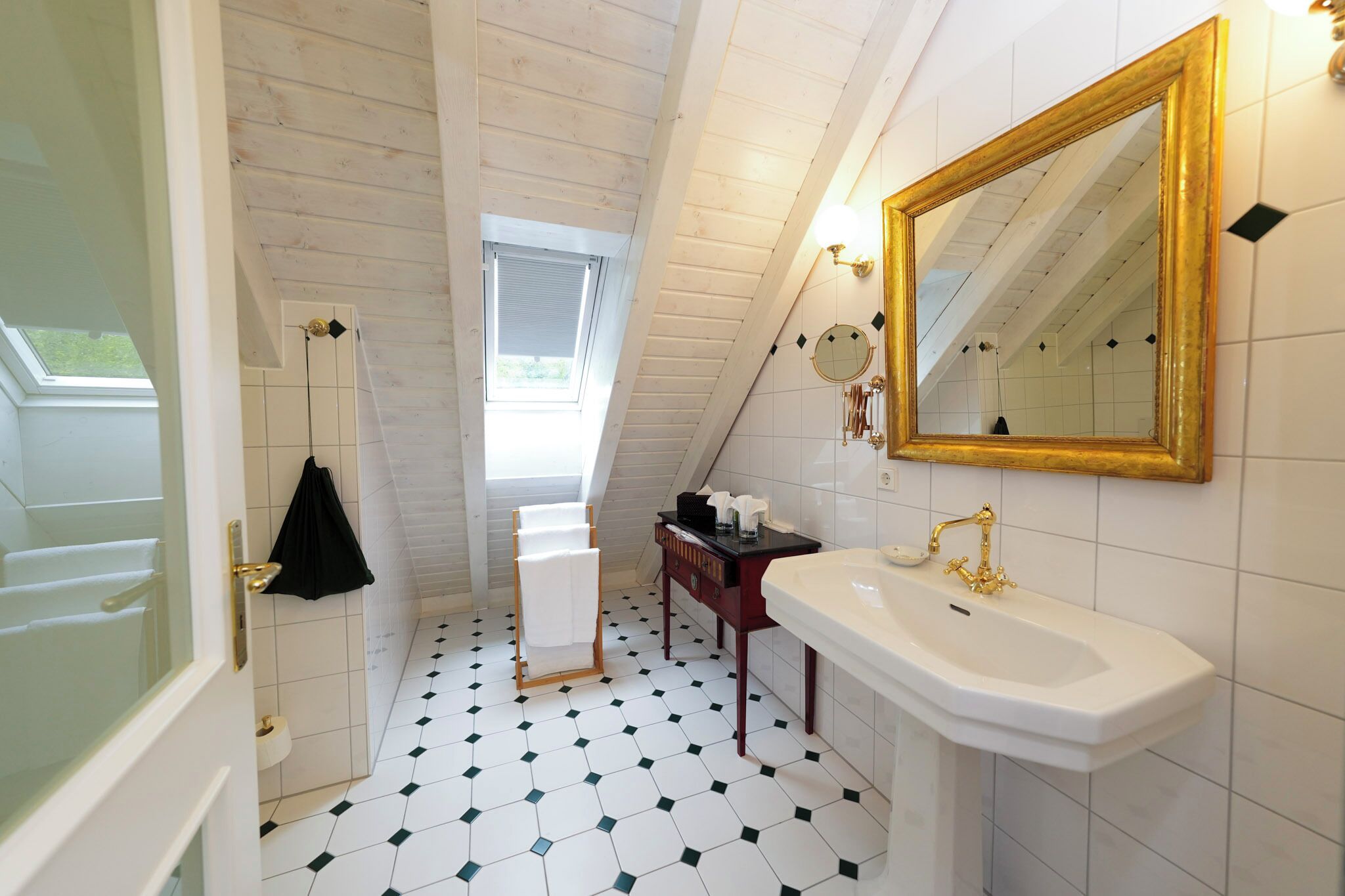 Une salle de bain avec un plafond en pente, un lavabo blanc avec des robinetteries dorées, un miroir avec un cadre doré et une serviette noire suspendue à un porte-serviettes.