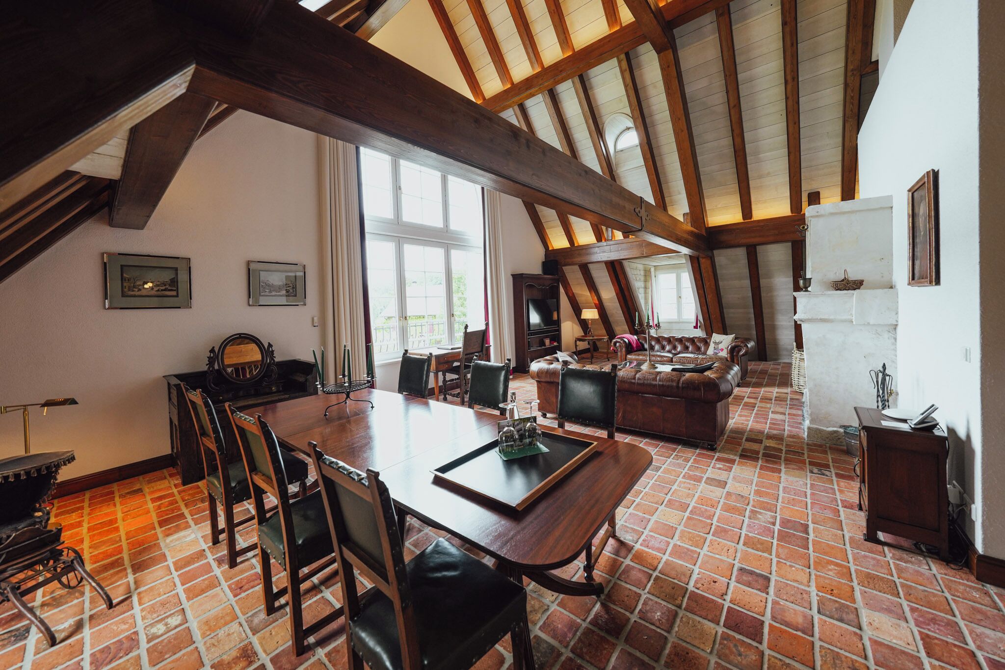 A spacious loft with a high ceiling, exposed wooden beams, a dining area with a long table and chairs, a cozy seating area with a sofa, and a kitchen area with a refrigerator and cabinets.