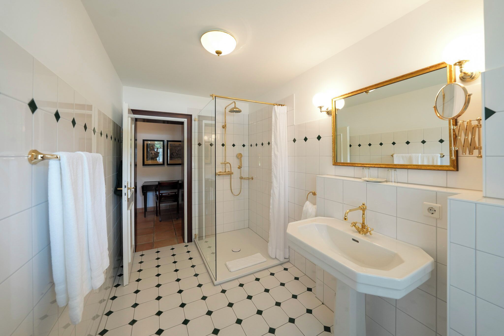 Ein Badezimmer mit einer gläsernen Duschkabine, einem weißen Waschbecken mit goldfarbenen Armaturen und einem großen Spiegel mit goldener Rahmung.