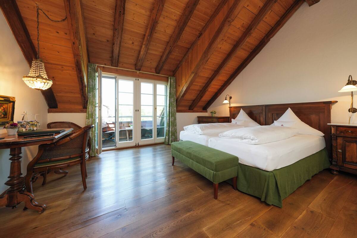 Una habitación con techo de madera, una cama con ropa de cama verde, un escritorio con una silla y una ventana con cortinas.
