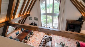 Una habitación con techos altos y vigas de madera a la vista, un amplio ventanal de arco, una chimenea y un área de comedor con mesa y sillas.