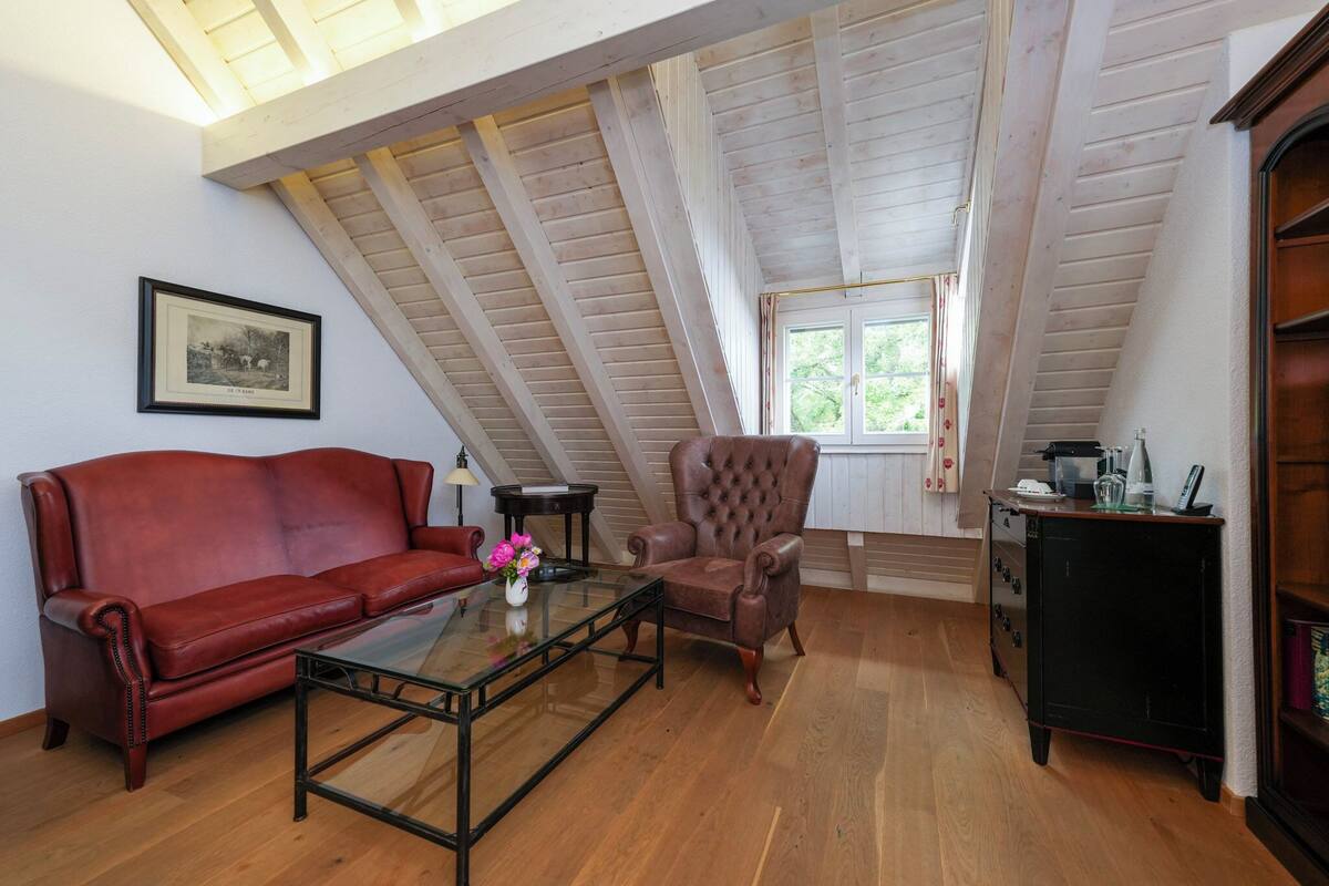 Un dormitorio tipo loft acogedor con techo inclinado, un sofá de cuero rojo, un sillón capitonado, una mesita con un florero y un aparador.
