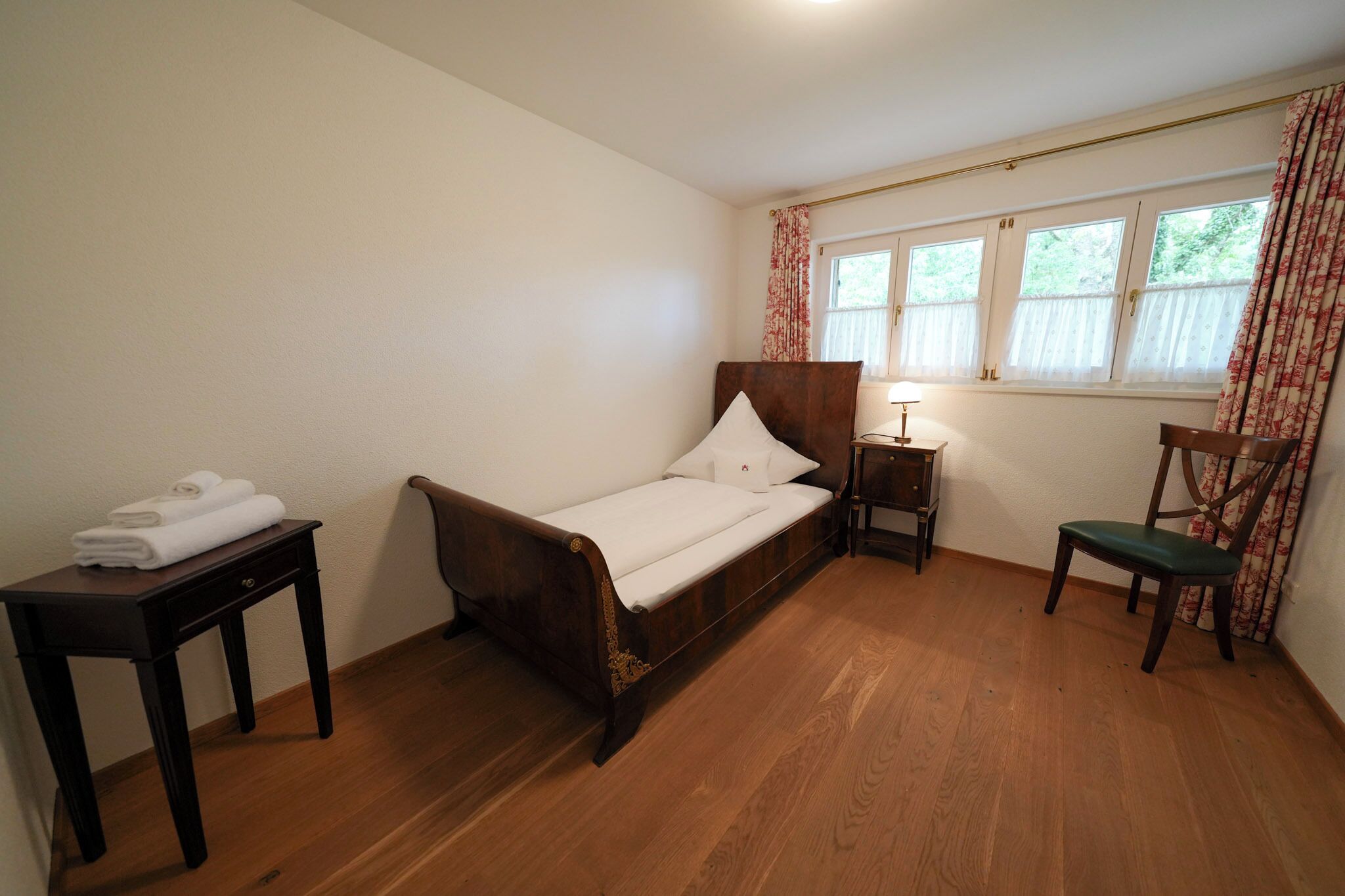 Ein kleines, ordentlich eingerichtetes Schlafzimmer mit einem Einzelbett, einem Nachttisch, einem Stuhl und einem Fenster mit Vorhängen.