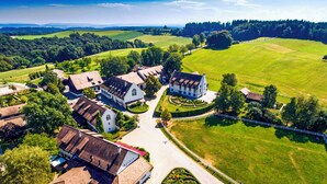 Vista aérea de una finca rural con varias construcciones, incluyendo una casa blanca grande con jardín, rodeada de extensos campos verdes y un área boscosa al fondo.