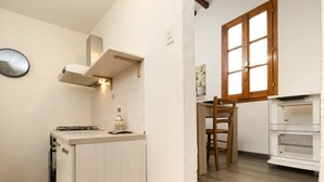 A compact kitchen with a gas stove, a built-in oven, and a white cabinet. There is a wooden chair and a small table with a plant. A window with wooden shutters is visible. - Santo Spirito Urban Apartment (Florence)