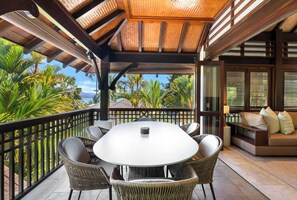 A covered outdoor area with a round table and chairs, a sofa, and a view of palm trees and the ocean.