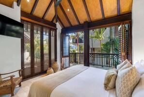 A bedroom with a large bed, wooden blinds, a TV, and a balcony with a view of palm trees.