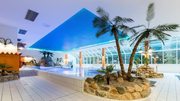 A modern hotel lobby with a spacious indoor pool, palm trees, and a glass-enclosed area.