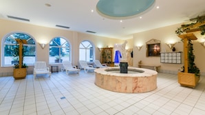 A spacious indoor area with a central circular marble fountain, white lounge chairs, and large windows providing natural light.