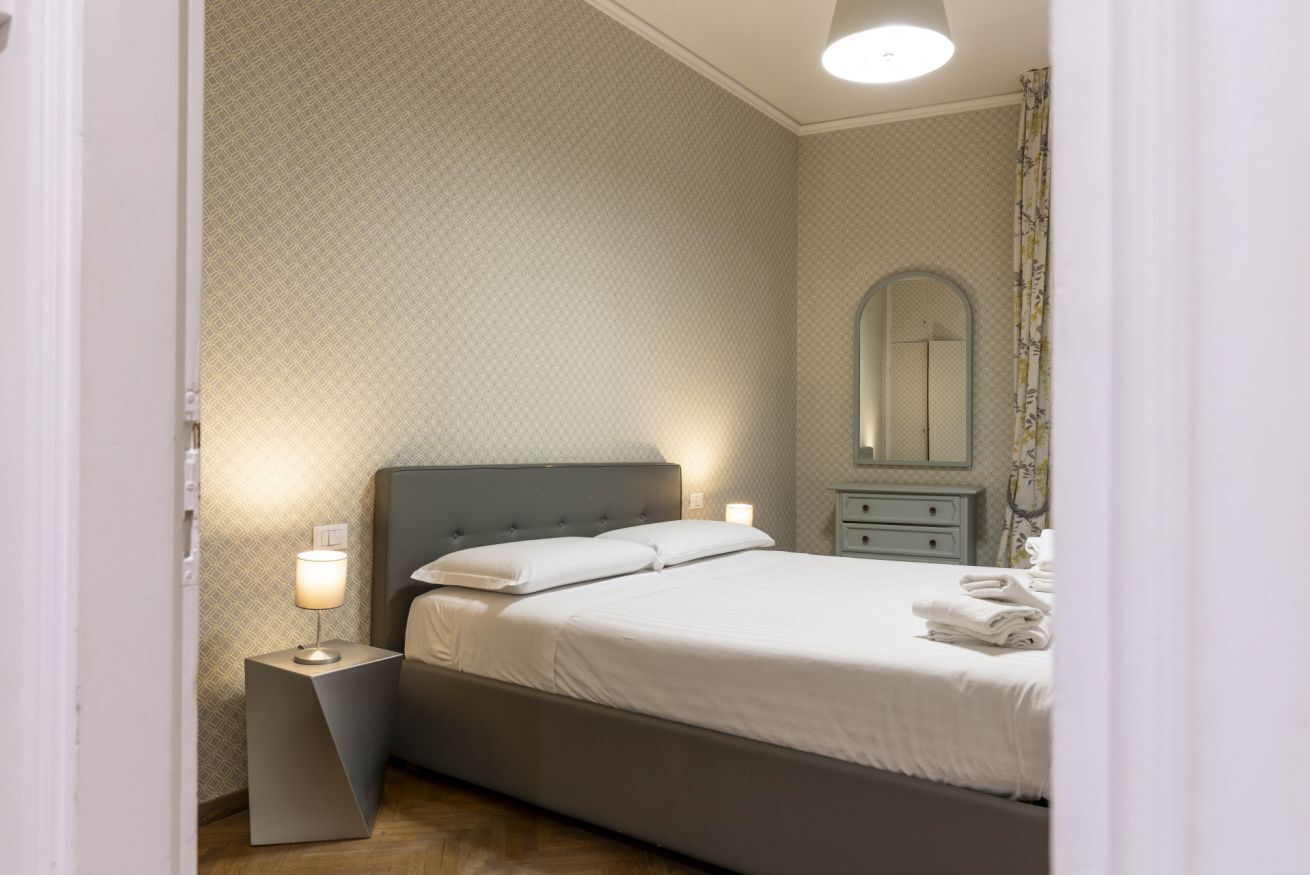 Un lit bien fait, avec du linge de lit blanc, une table de chevet avec une lampe et une petite commode avec un miroir.