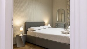 Un letto rifatto con lenzuola bianche, un comodino con una lampada e un piccolo settimino con uno specchio.