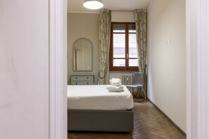 Un dormitorio con cama, cómoda, espejo, radiador y ventana con cortinas.