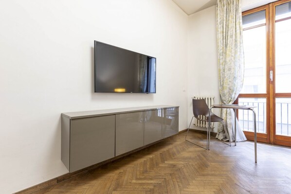 Una sala de estar moderna con un televisor de pantalla plana en la pared, un mueble gris, piso de madera con patrón de espiga, una mesa de comedor con una silla y un ventanal con cortinas.