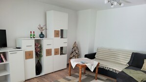 A modern living room with a wooden floor, a black sofa, a white cabinet with decorative panels, a small wooden table, and a television on a stand. - Cozy newly built vacation apartment in quiet wooded surroundings (Dippoldiswalde)