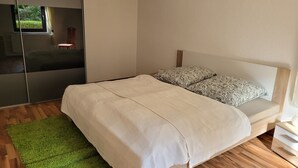 A bedroom with a bed, a green rug, a wooden floor, a window, and a mirror. - Cozy newly built vacation apartment in quiet wooded surroundings (Dippoldiswalde)