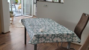 A dining area with a table covered by a floral tablecloth, a brown upholstered chair, and a view of a kitchen and outdoor area through a glass door. - Cozy newly built vacation apartment in quiet wooded surroundings (Dippoldiswalde)