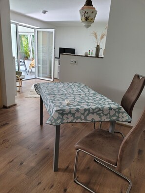A dining area with a table covered by a floral tablecloth, a brown upholstered chair, and a view of a kitchen and outdoor area through a glass door.
