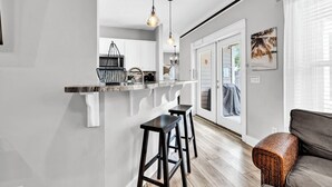 Une cuisine moderne avec des armoires blanches, un comptoir noir et un luminaire suspendu. Le sol est en bois et il y a un canapé gris dans l’espace salon.