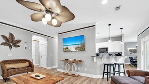 Un salon moderne avec une table basse en bois, un grand ventilateur de plafond et un coin repas équipé de tabourets de bar.