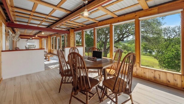 A spacious room with a large dining table and chairs, wooden beams on the ceiling, and large windows offering a view of the outdoors. - Lakefront Retreat: Private Dock | King Bedrooms (Burnet County)