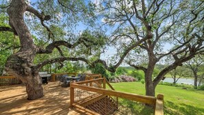 A wooden deck with outdoor furniture, surrounded by large trees and a view of a body of water. - Lakefront Retreat: Private Dock | King Bedrooms (Burnet County)