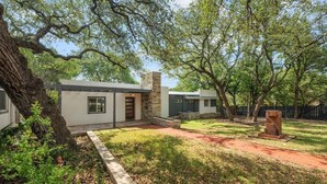 A modern two-story house with a stone fireplace, surrounded by a well-maintained lawn and mature trees. - Lakefront Retreat: Private Dock | King Bedrooms (Burnet County)