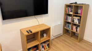 A room with a flat-screen TV mounted on the wall, a wooden bookshelf filled with books, and a small wooden table with a remote control and a mug. - Deluxe Twin Room with Shared Kitchen & Bathroom Access - For 2 Guests (London)