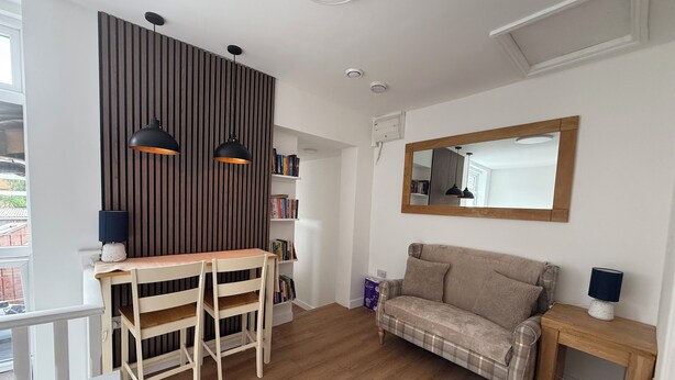 A modern living room with a wooden floor, a sofa, a dining table with chairs, a bookshelf, and two pendant lights. - Deluxe Twin Room with Shared Kitchen & Bathroom Access - For 2 Guests (London)