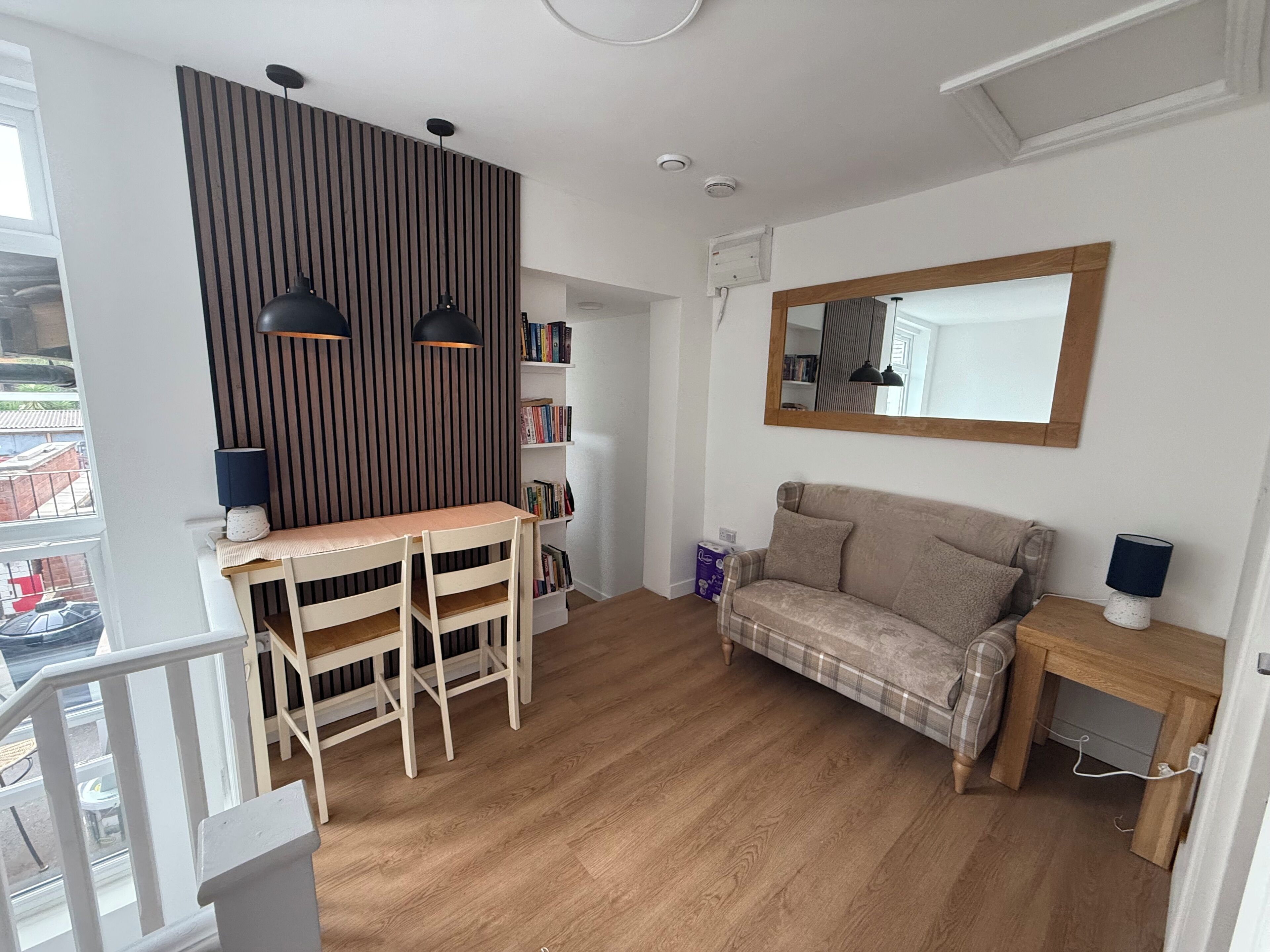 A modern living room with a wooden floor, a sofa, a dining table with chairs, a bookshelf, and a mirror on the wall.