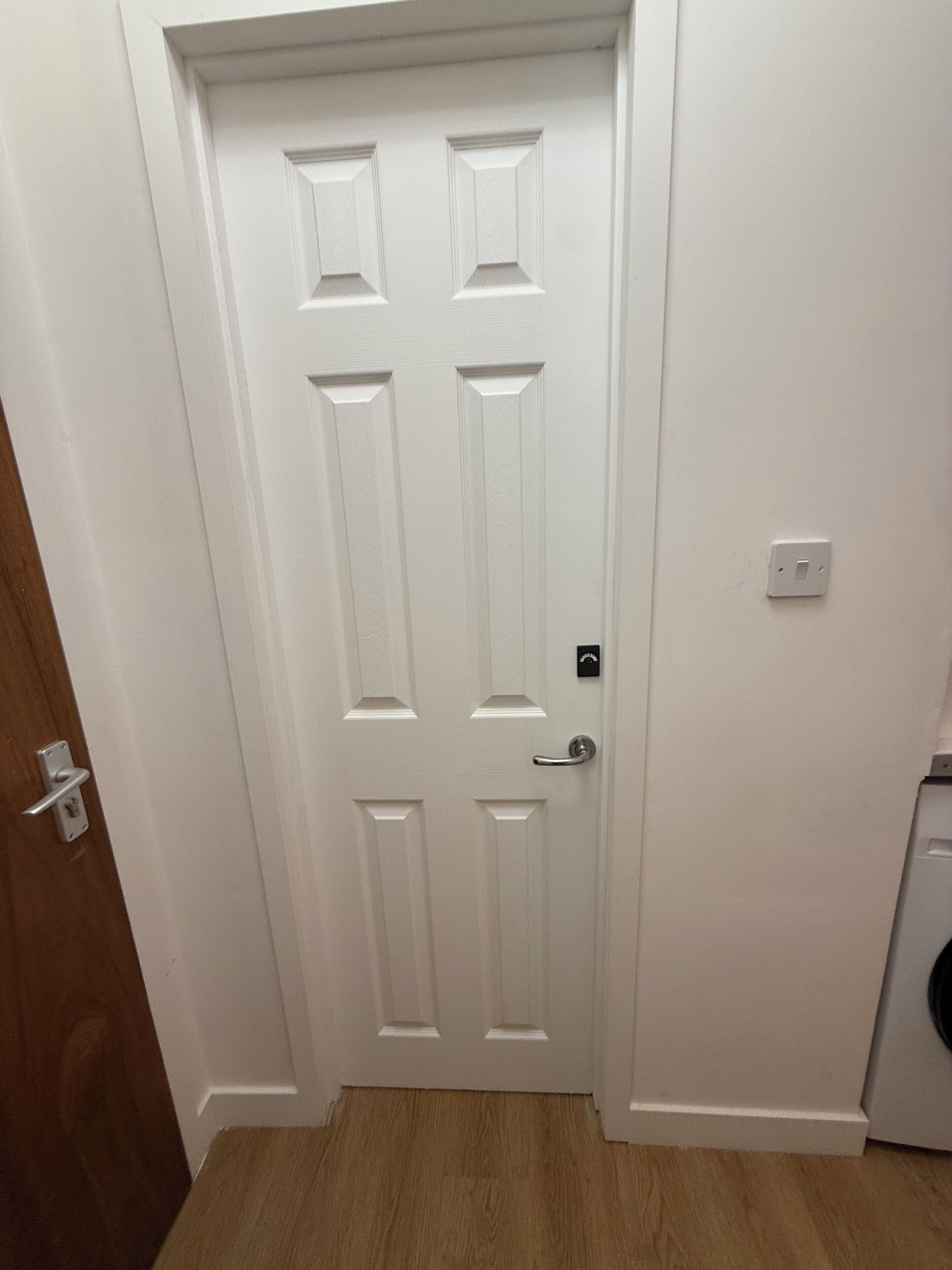 A white door with a silver handle and a keyhole, a light switch on the wall, and a wooden floor.