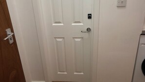 A white door with a silver handle and a keyhole, a light switch on the wall, and a wooden floor. - Deluxe Twin Room with Shared Kitchen & Bathroom Access - For 2 Guests (London)