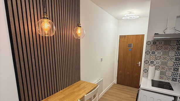 A compact kitchen with a wooden table and chairs, a built-in stove, and a wall-mounted light fixture. - Modern Studio with private Bathroom & Kitchenette in 3 bed apartment (London)