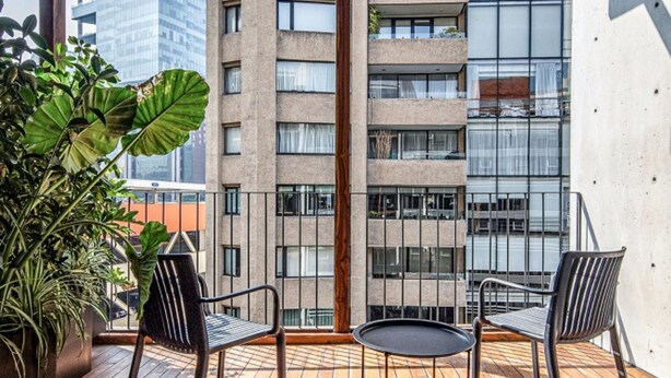 Een houten terras met twee stoelen en een tafel, met uitzicht op een modern gebouw met balkons.
